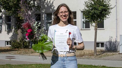 Die Preisträgerin steht auf der Wiese vor einem Hochschulgebäude und hält lachend ihre Urkunde sowie den Blumenstrauß in der Hand.