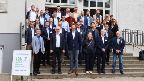 Gruppenfoto von etwa 30 Teilnehmer_innen der 106. BundesDekaneKonferenz Wirtschaft an der Hochschule Wismar. Die Personen stehen auf einer Außentreppe vor einem hellen Gebäude mit großen Fenstern. Links im Vordergrund ist ein Schild mit der Aufschrift „Herzlich Willkommen 106. BundesDekaneKonferenz – Transfer und Wandel“ zu sehen.