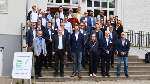 Gruppenfoto von etwa 30 Teilnehmer_innen der 106. BundesDekaneKonferenz Wirtschaft an der Hochschule Wismar. Die Personen stehen auf einer Außentreppe vor einem hellen Gebäude mit großen Fenstern. Links im Vordergrund ist ein Schild mit der Aufschrift „Herzlich Willkommen 106. BundesDekaneKonferenz – Transfer und Wandel“ zu sehen.