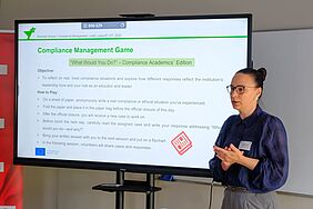A woman speaks in front of a large screen displaying the “Compliance Management Game” instructions. The game involves real-life ethical scenarios in teaching contexts.