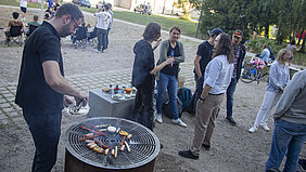 Eine Person steht an einem Grill auf dem verschiedene Speisen liegen. Dahinter stehen Personen, die sich unterhalten.