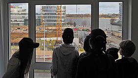 Es sind sechs Personen als Schattenriss vor einem Fenster zu sehen, die auf eine Baustelle blicken.