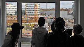 Es sind sechs Personen als Schattenriss vor einem Fenster zu sehen, die auf eine Baustelle blicken.