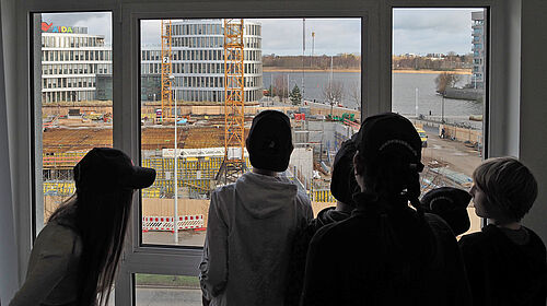 Es sind sechs Personen als Schattenriss vor einem Fenster zu sehen, die auf eine Baustelle blicken.