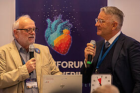 Noelle-Pawlak A: Die beiden mit Sakkos bekleideten Männer haben Mikrofone in der Hand und schauen sich an. Im Hintergrund befindet sich ein Banner mit einer Herzgrafik.