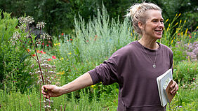 Eine Frau steht in einem blühenden Schaugarten. Sie hält in einer Hand eine Pflanze und in der anderen ein Notizbuch