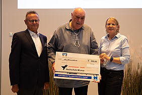 Drei Personen stehen nebeneinander in einem Raum. Die mittlere hält einen großen Scheck mit der Aufschrift "Hochschule Wismar" und "300,00 Euro". Alle lächeln.