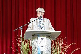 Mann mit Brille, Sakko und einer großen Amtskette steht am Rednerpult vor rotem Theatervorhang und spricht in Mikrofon