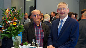 Die beiden genannten Personen stehen an einem Tisch, auf dem sich zwei Sektgläser und ein großer Blumenstrauß befinden. Im Hintergrund sind zahlreiche Gäste zu sehen.