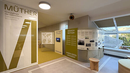 An exhibition space with information panels and a screen displaying the exhibition on Müther at the Hyparforum, with seating and large windows in the background.