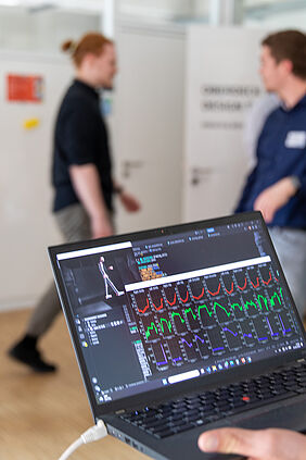 Es ist der Bildschirm eines Laptops zu sehen. In dessen linker oberer Ecke ist eine Art Strichmännchen zu erkennen. Im Hintergrund bewegt sich die Person, deren Gang gemessen wird. Sie ist unscharf.