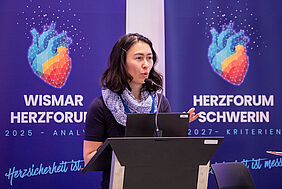 Prof. Schmolke Die Professorin steht hinter einem Pult vor zwei farbigen Bannern, auf denen eine Herzgrafik zu sehen ist. Sie blickt in Richtung Publikum.