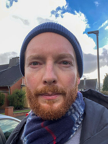 Die Person ist im Porträt zu sehen. Sie steht im Freien, hat eine Mütze auf und einen Schal um.