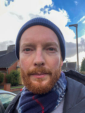 Die Person ist im Porträt zu sehen. Sie steht im Freien, hat eine Mütze auf und einen Schal um.