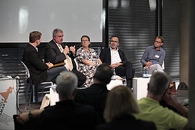 Der Blick geht über zwei Publikumsreiehn hinweg Richtung Podium. Dort sitzen fünf Personen. Ein Mann gestikuliert.