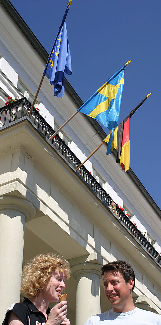 Zwei lachende Personen im Sommer vor einem Gebäude mit verschiedenen Flaggen